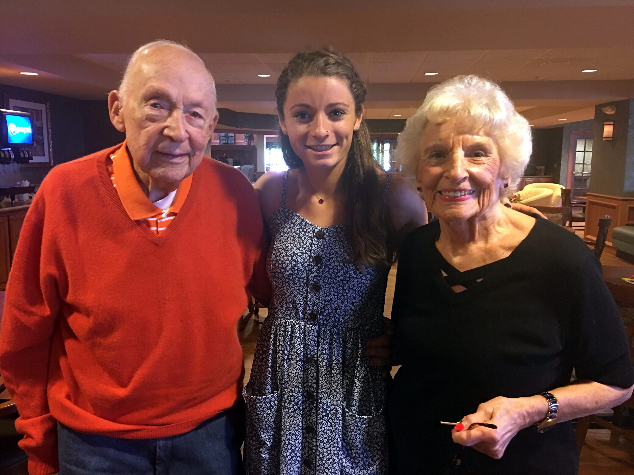 Three smiling adults pose closely together indoors: an older man in a red sweater on the left, a younger woman in the middle, and an older woman in black on the right.
