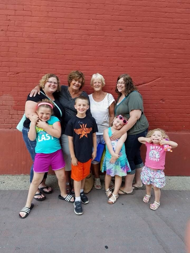 Group of adults and children smiling while posing together in front of a red brick wall on a sidewalk.