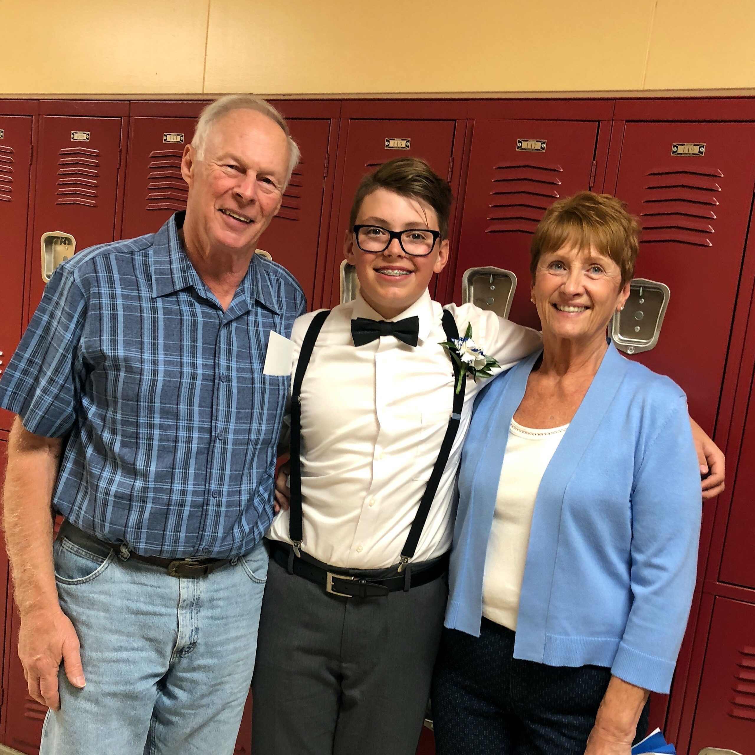 Three people posing in front of red lockers: an older man in a plaid shirt, a teen with glasses and a bow tie, and a woman in a light blue blazer.