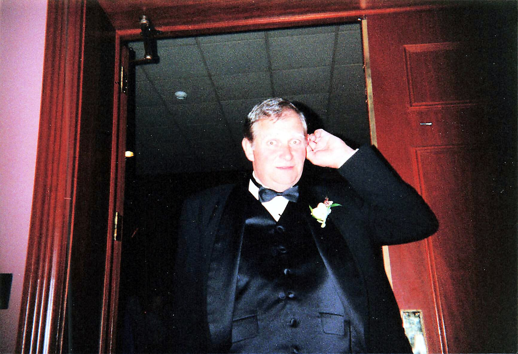 Man in a black tuxedo with bow tie and boutonniere adjusting his earpiece at a wooden doorway during a formal event.