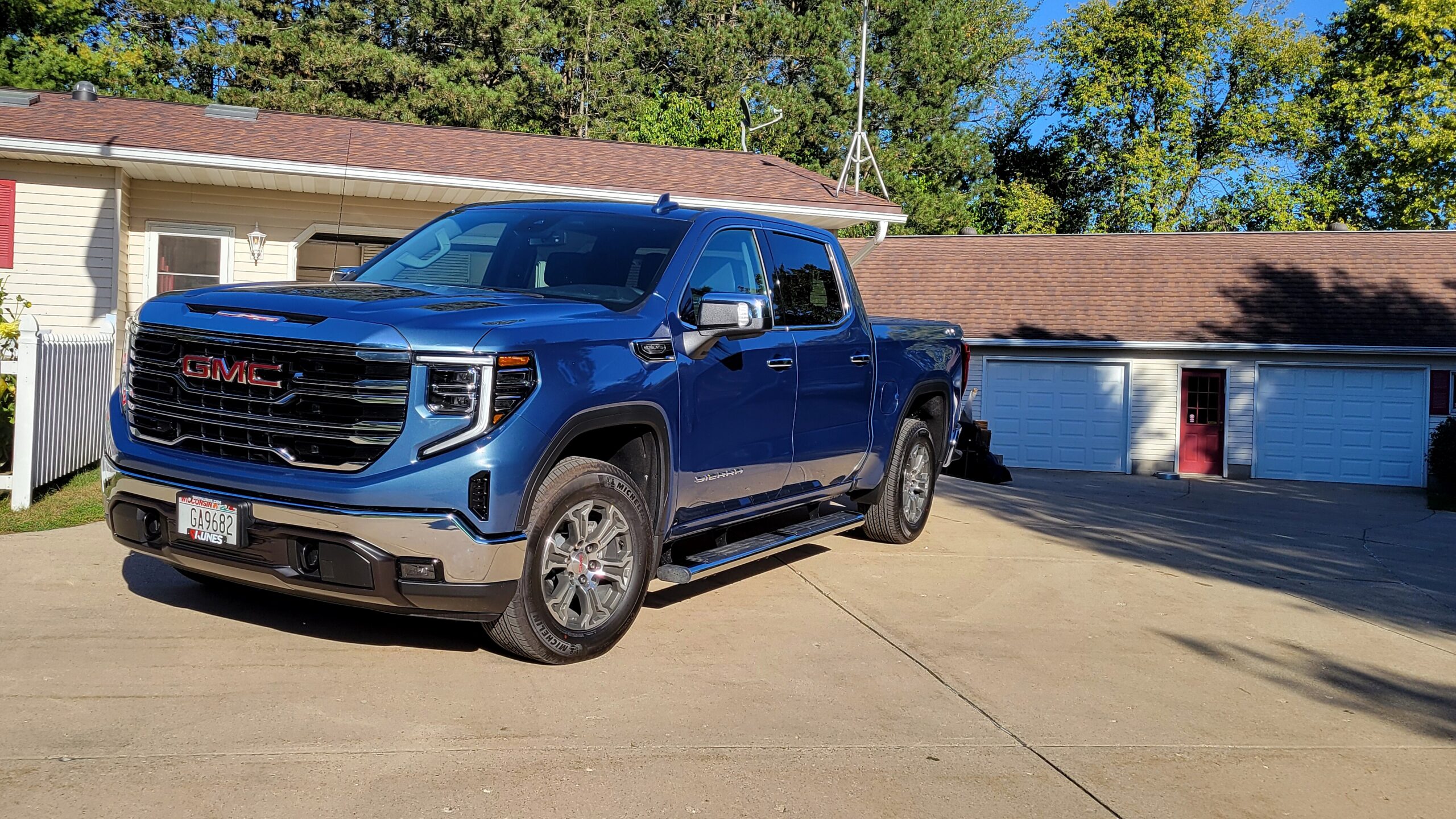 Blue GMC Sierra pickup parked in a driveway in front of a suburban house with a two-car garage and red front door behind it.