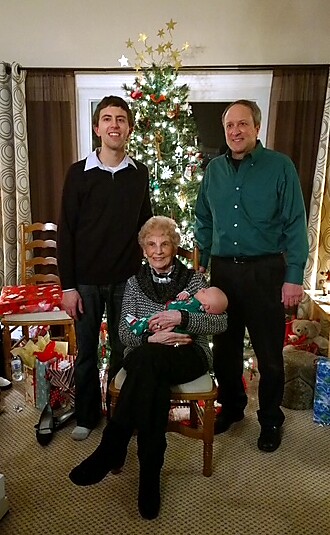 Three generations posing for a Christmas photo: an elderly woman seated holding a baby, with two men standing beside her near a decorated Christmas tree.