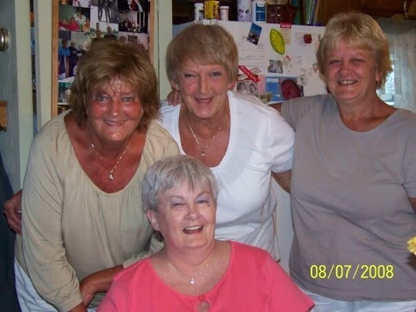 Four women smiling together in a home kitchen, one seated in front wearing pink.