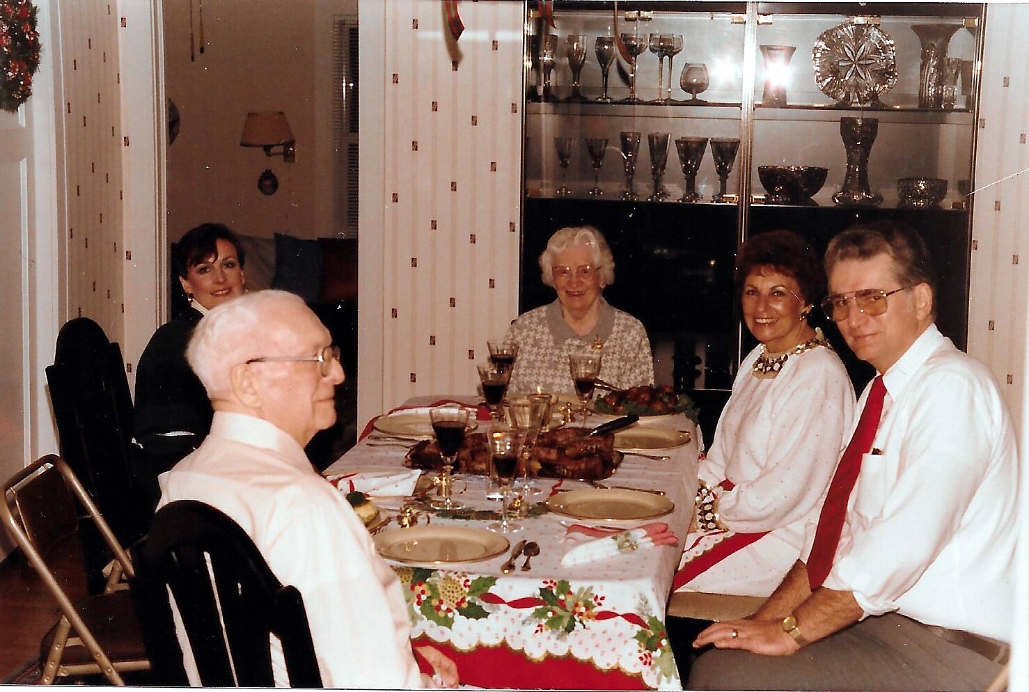 Six people seated at a Christmas dinner table with a festive tablecloth, wine glasses, and a glass-front cabinet in the background.