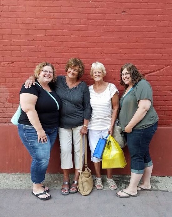 Four women standing arm in arm and smiling against a red brick wall, each with shopping bags.