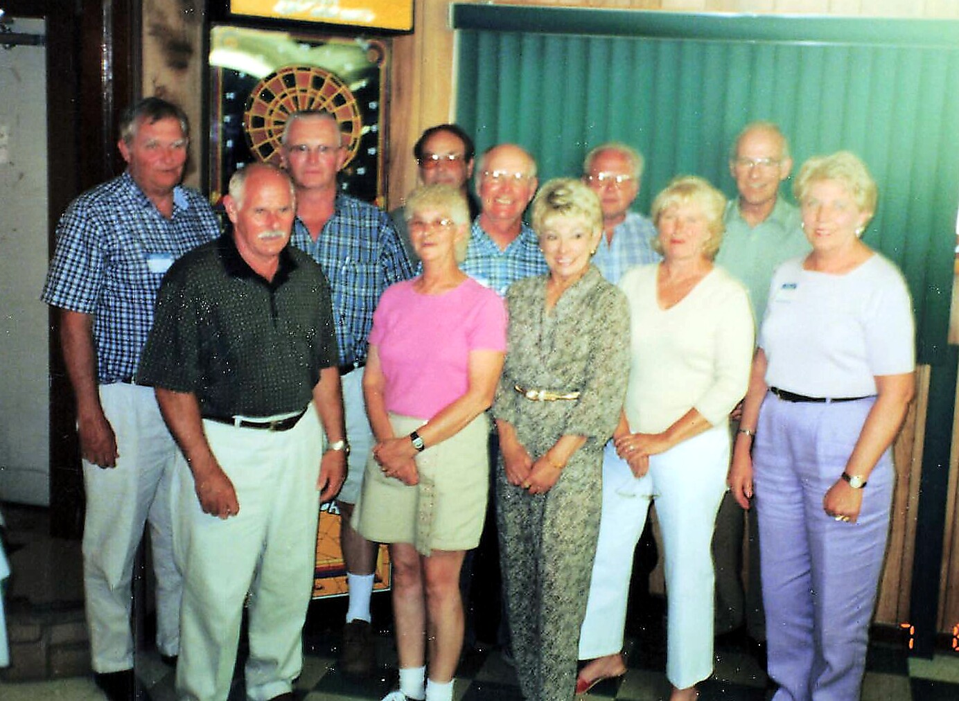 Group of older adults posing for a casual group photo indoors, with a dartboard in the background.