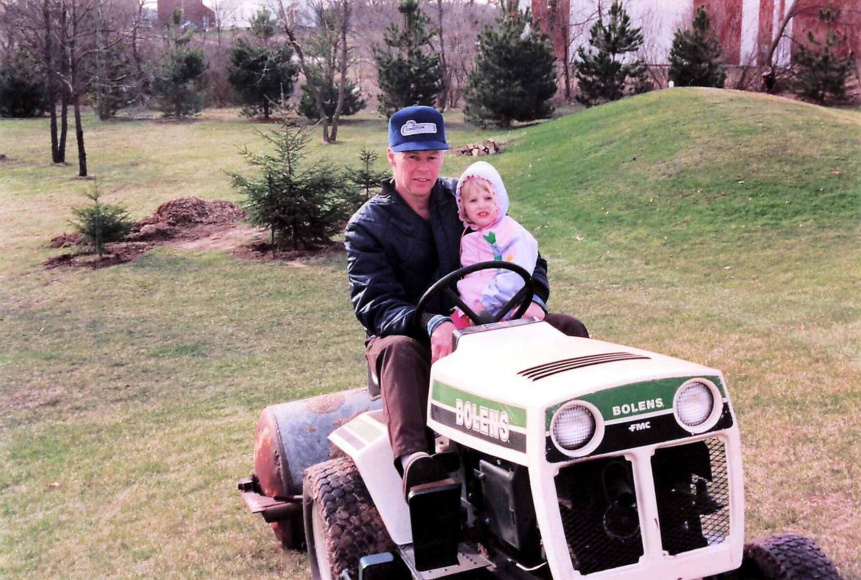 Man and child ride a white and green Bolens garden tractor across a grassy yard, the man steering while the child sits beside him in a hooded jacket.
