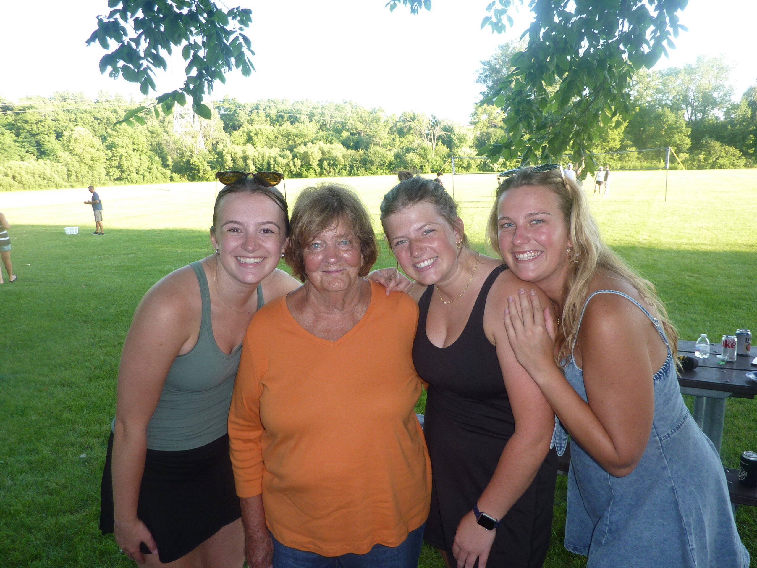 Four friends posing together in a sunny park, smiles for the camera with trees and grass in the background.