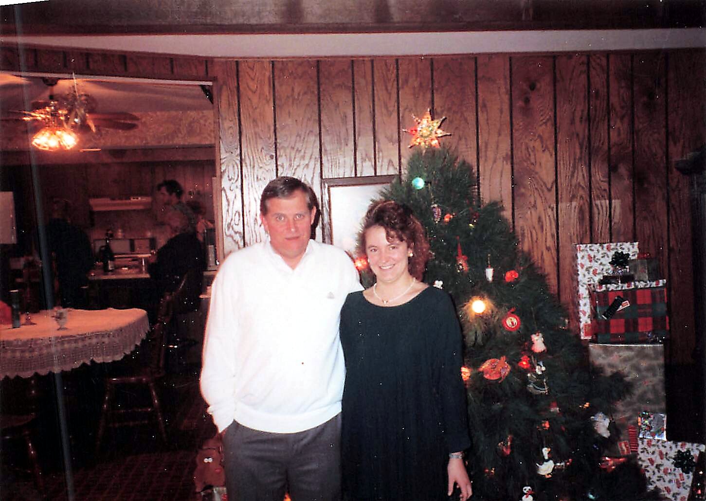 Man and woman stand together by a decorated Christmas tree with gifts underneath in a wood-paneled living room.