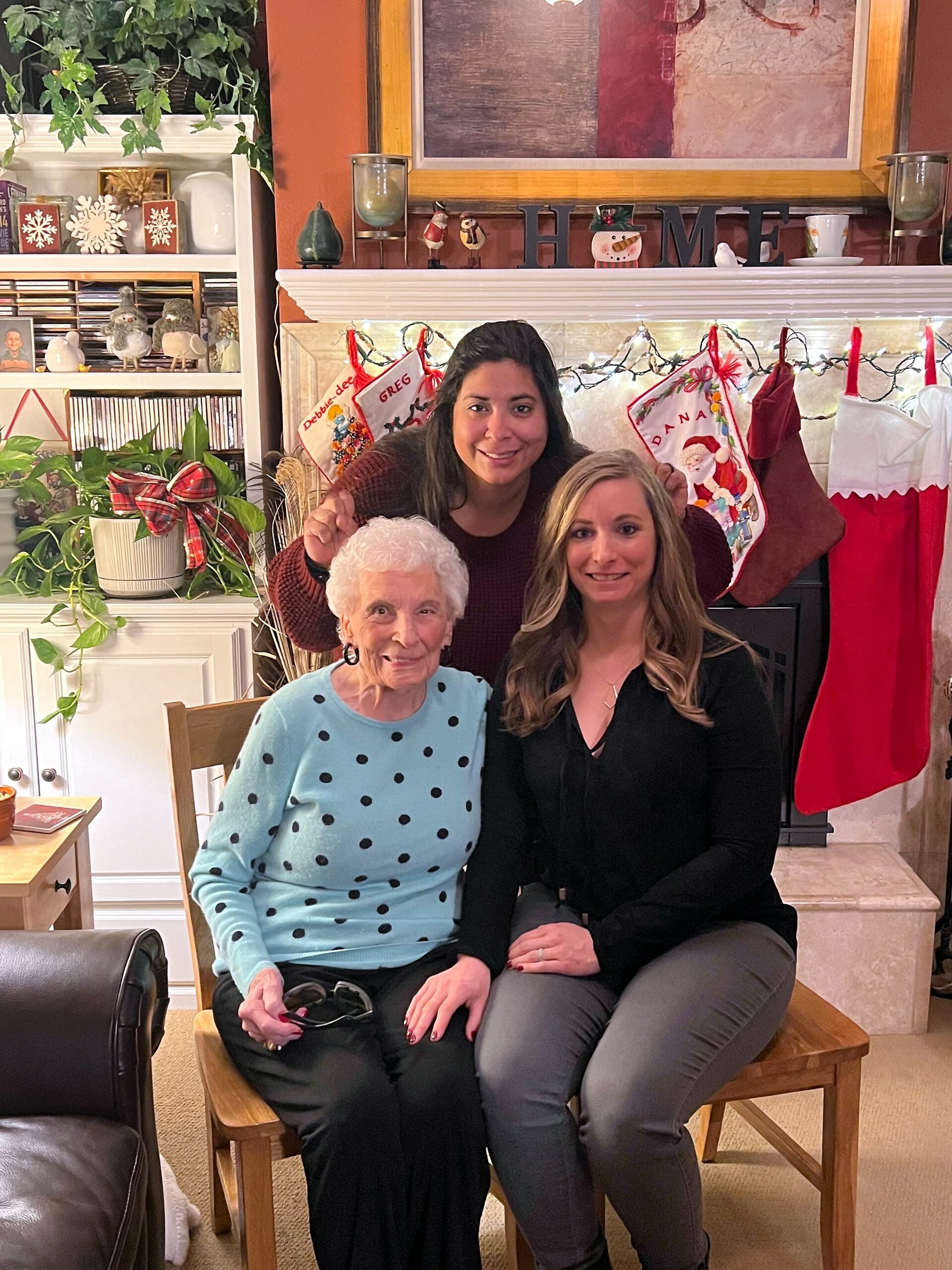 Three generations of women posing together in a festively decorated living room with stockings and Christmas lights (smiling).