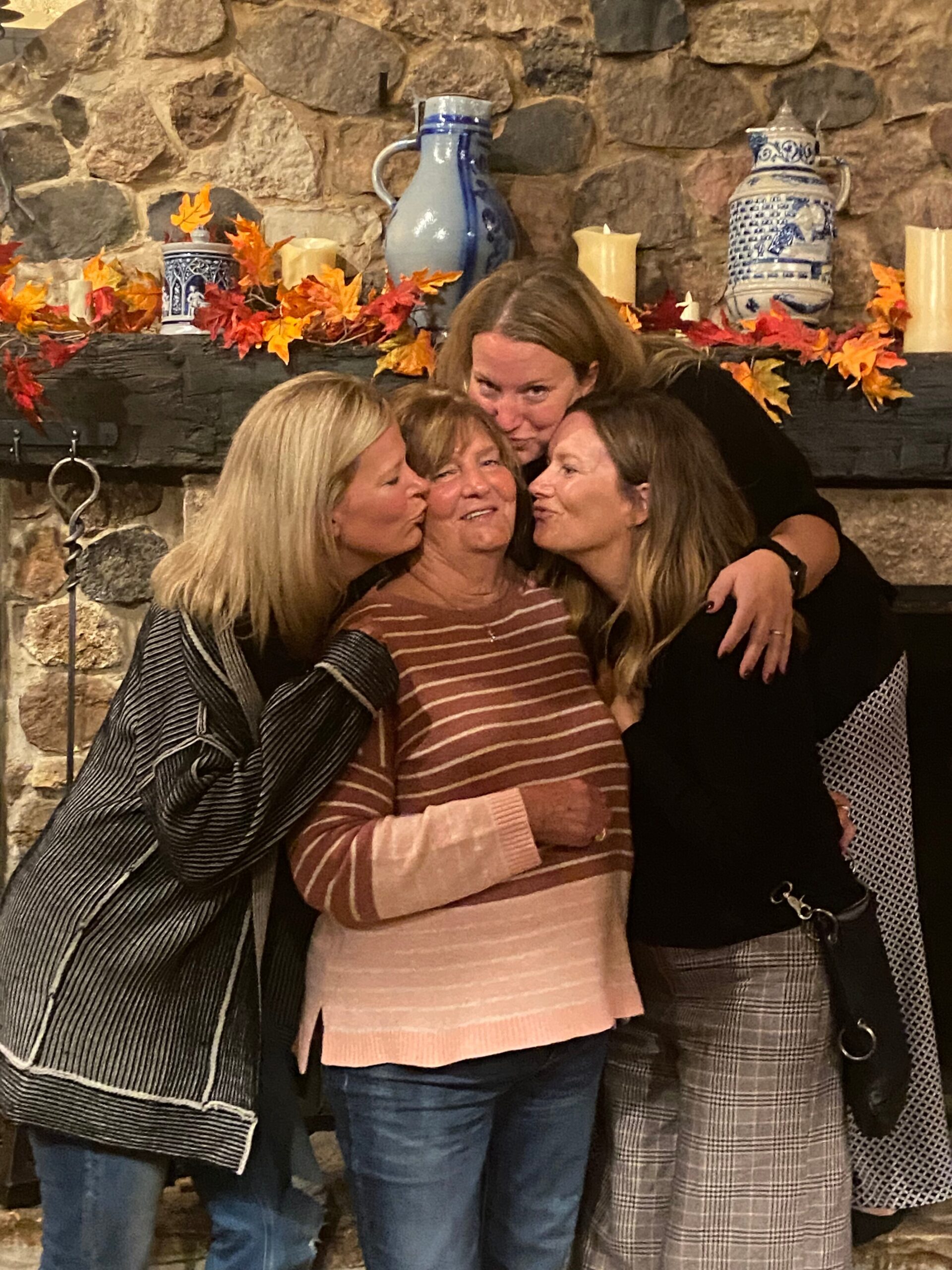 Four women hug and kiss an older woman on the cheek in front of a stone fireplace decorated with autumn leaves and blue ceramics.