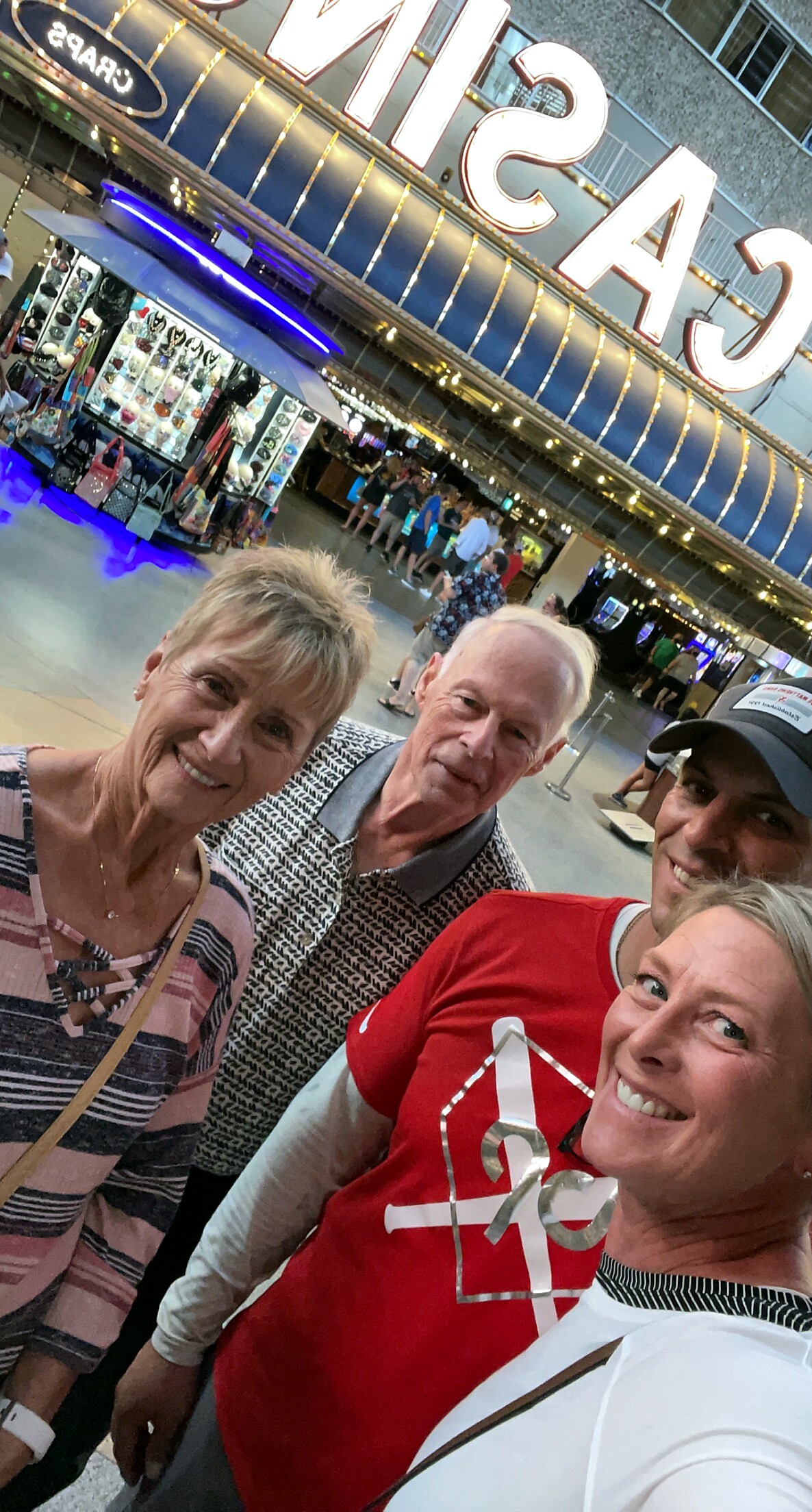 Four friends take a cheerful selfie inside a casino lobby, with bright signage and souvenir stalls in the background.