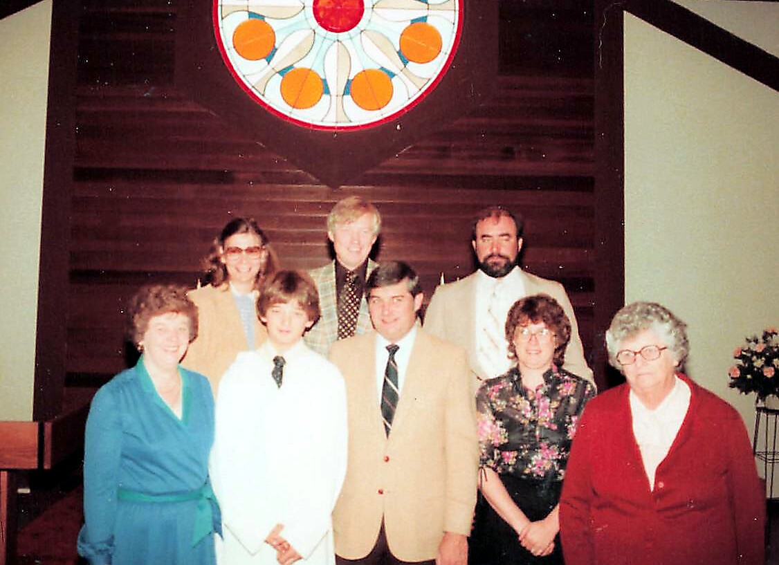 Group of eight people posing for a photo in a church, standing under a circular stained glass window with red, orange, and blue colors in the background.