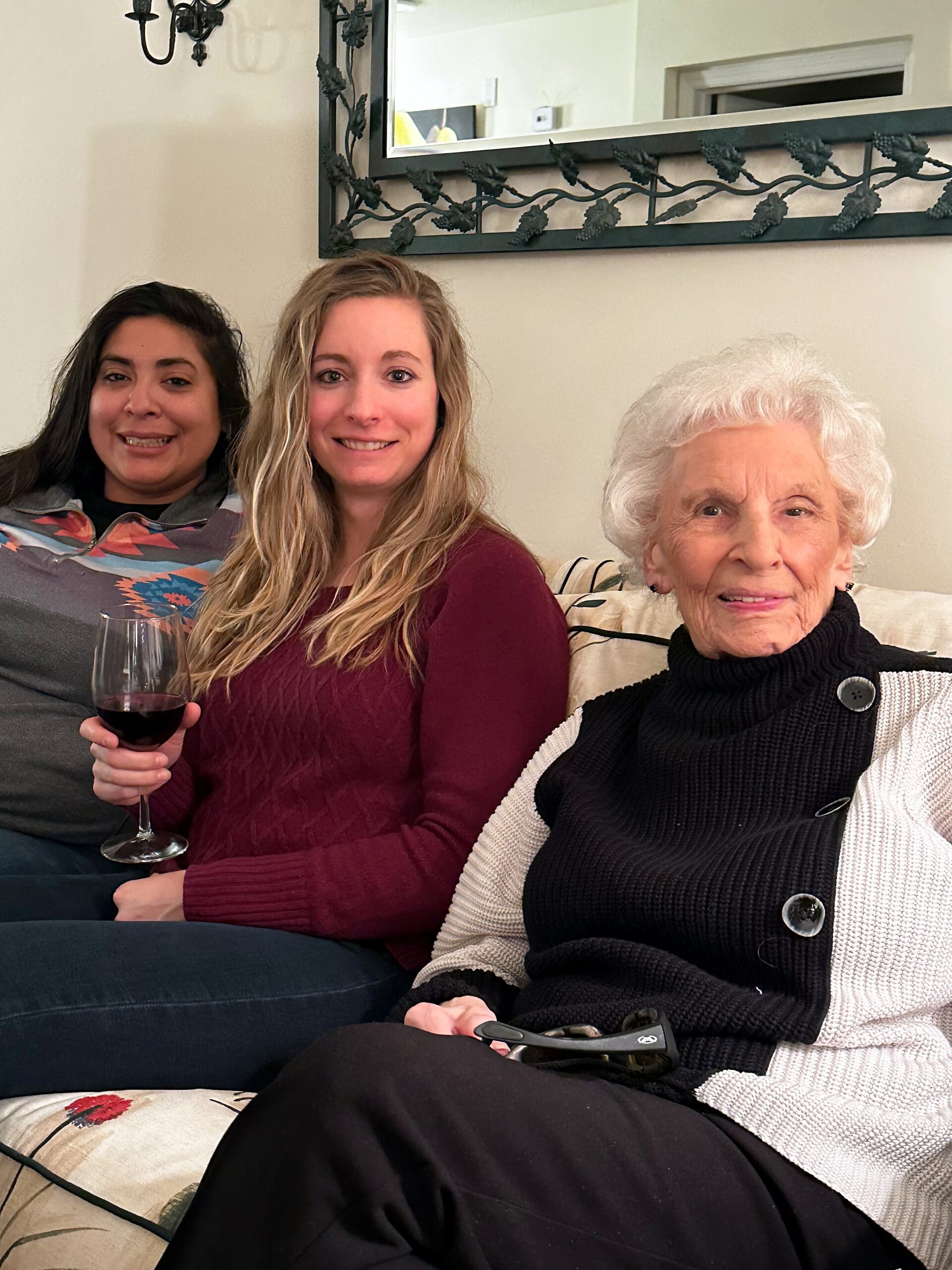 Three women with light skin sit on a couch, smiling; the leftmost woman holds a glass of red wine.