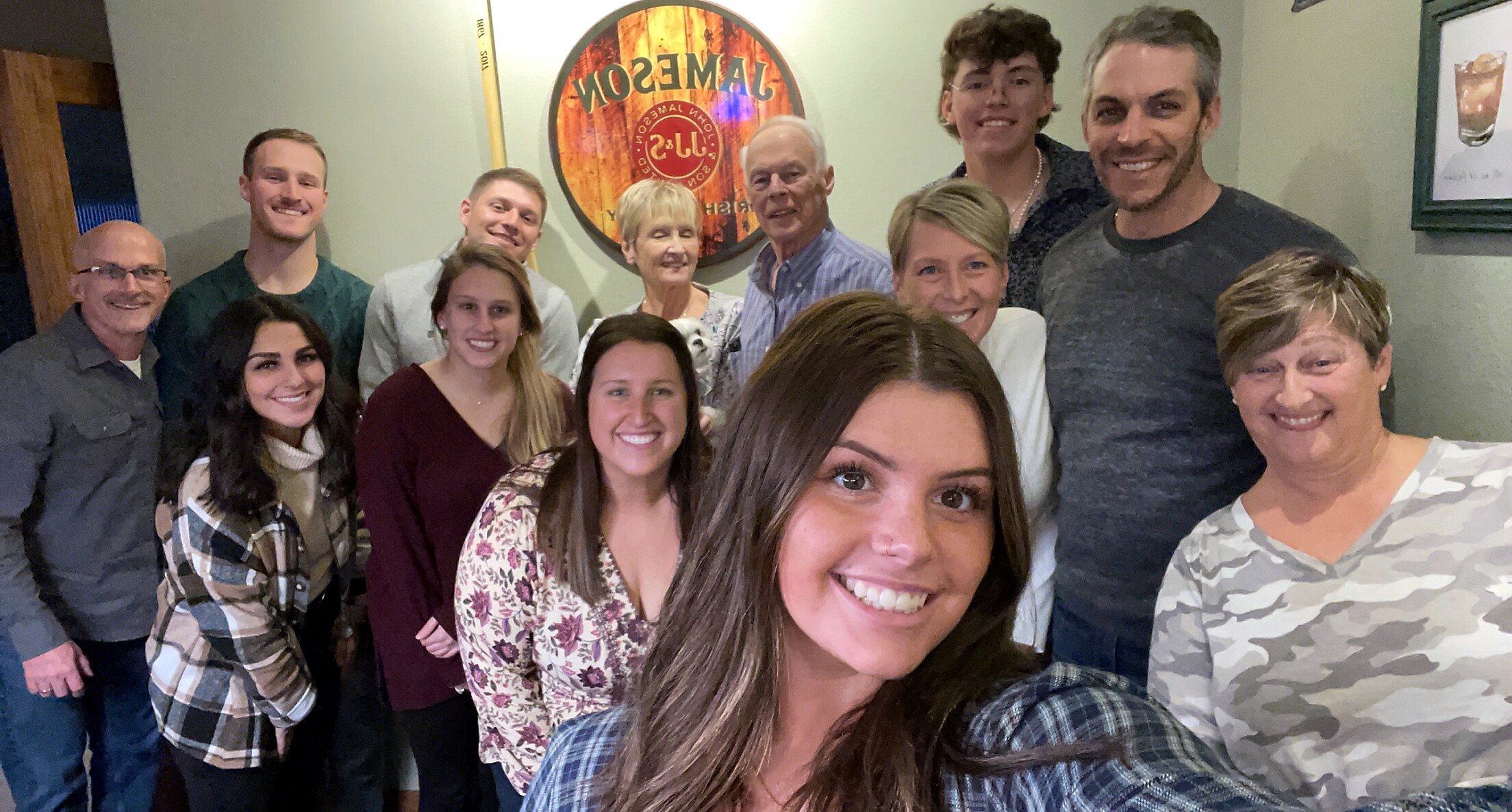 Twelve people pose for a group selfie in a cozy room, smiling at the camera, with a round wall sign in the background.