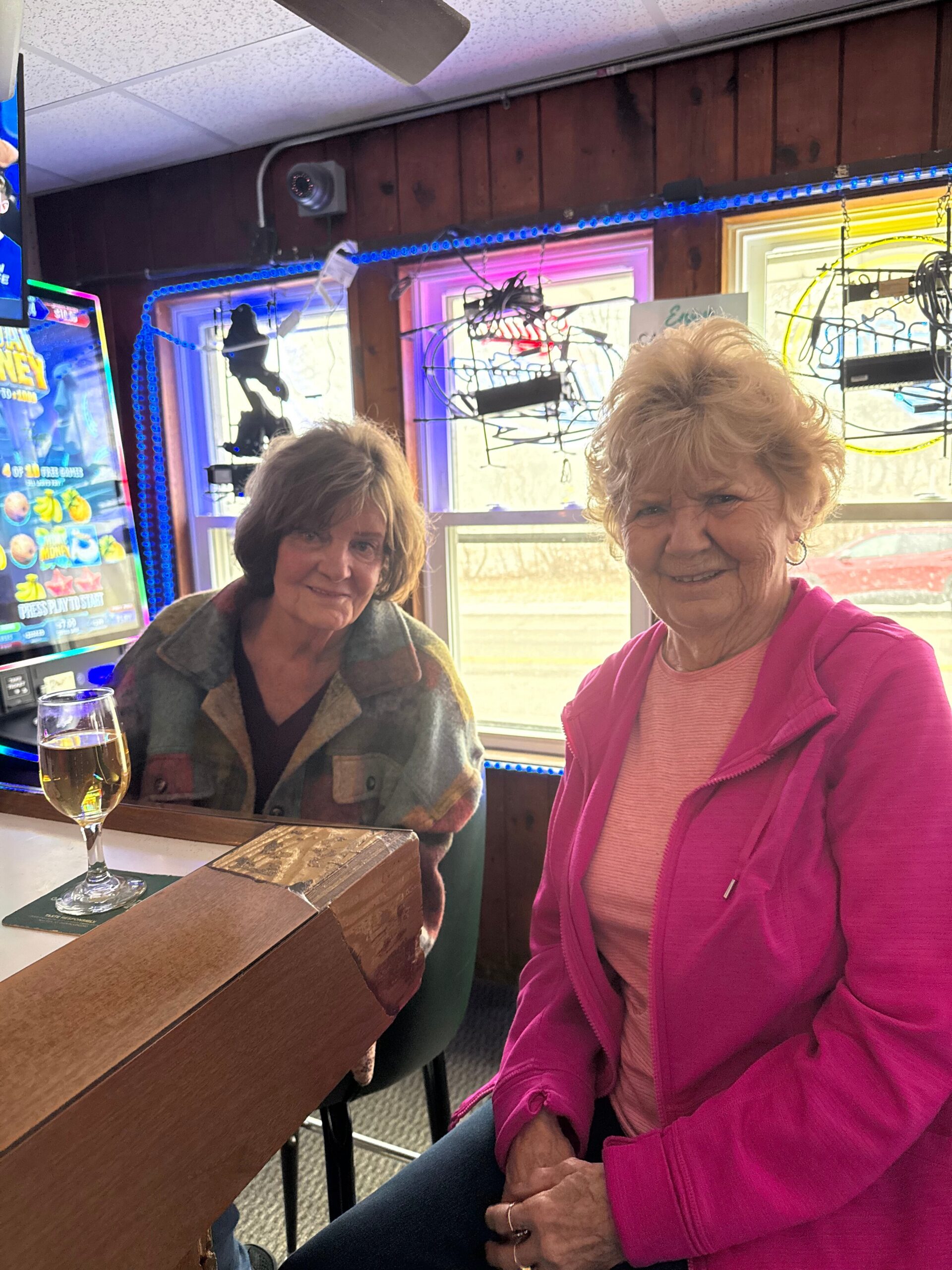 Two smiling older women sit at a wooden bar counter, one in a pink jacket, with a glass of beer nearby on the counter.