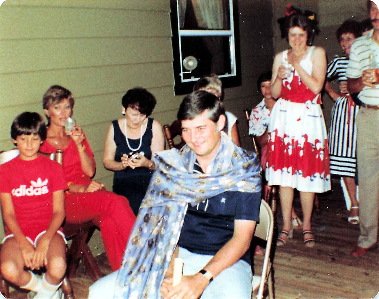 Group of adults at a summer porch party; man sits center wearing a floral scarf, others stand or sit nearby with drinks and a woman in a red dress visible in the background.