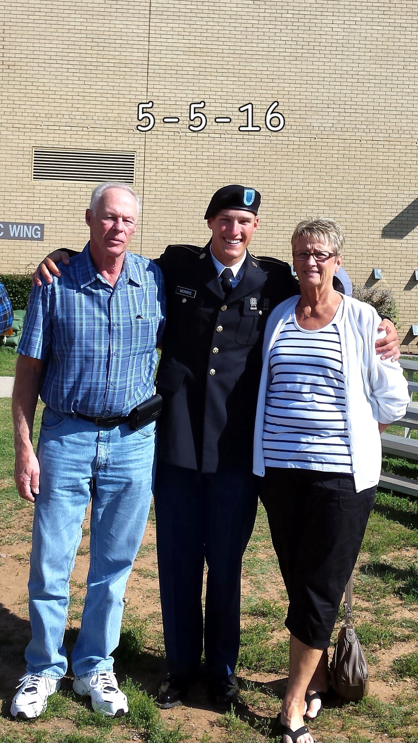 Three adults posing together outside a brick building; a man in blue plaid shirt, a smiling soldier in dress uniform, and a woman in striped top.
