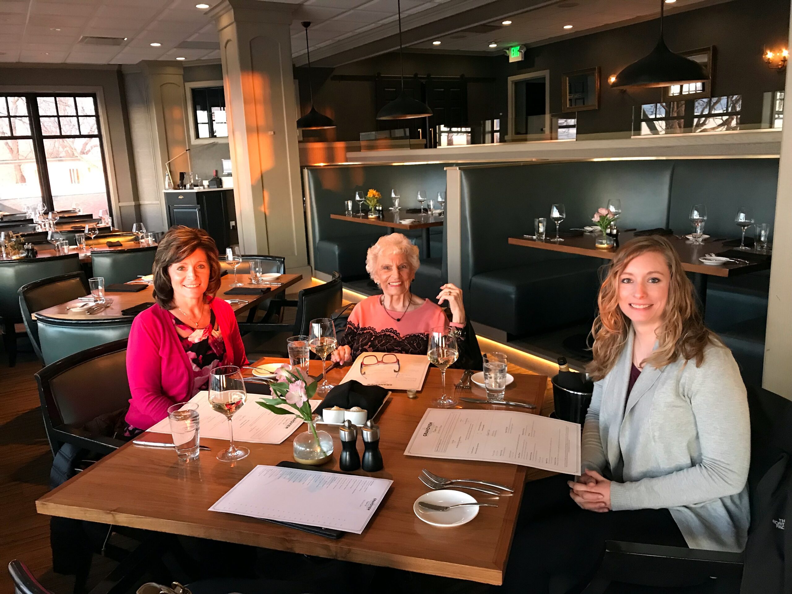 Three women sitting at a restaurant table with menus, glasses of wine, and a small flower centerpiece.