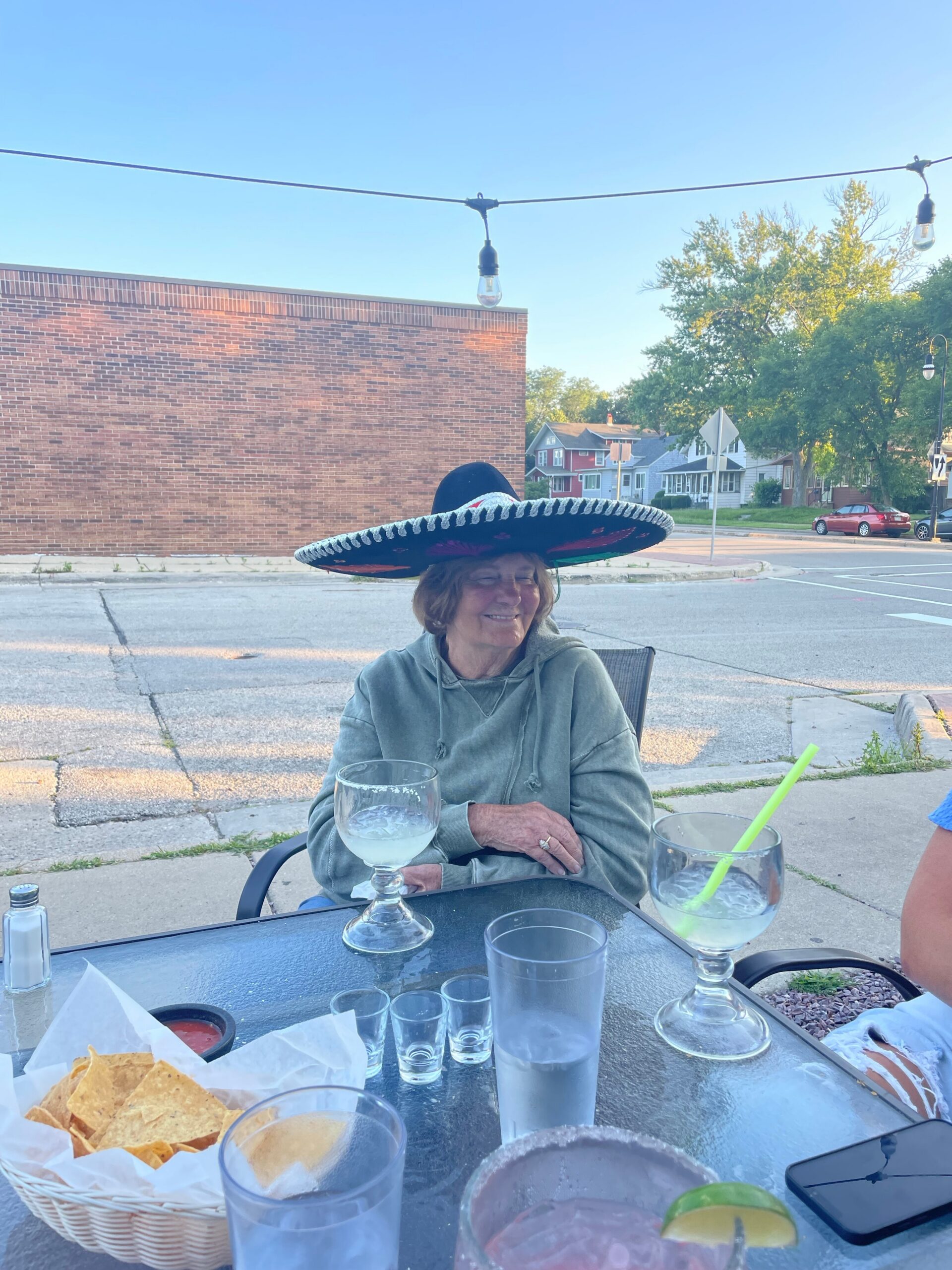 Older woman wearing a large sombrero sits at a glass outdoor table with drinks and tortilla chips, smiling at the camera.
