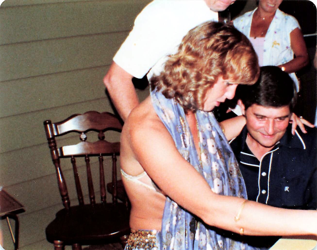 Woman with curly hair and a scarf leaning toward a smiling man seated at a casual porch party.