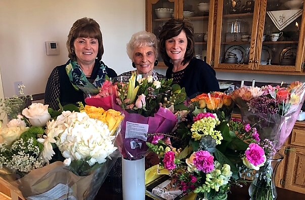 Three women in a home kitchen smile at the camera, standing behind a table full of vibrant bouquets including pinks, yellows, and whites.