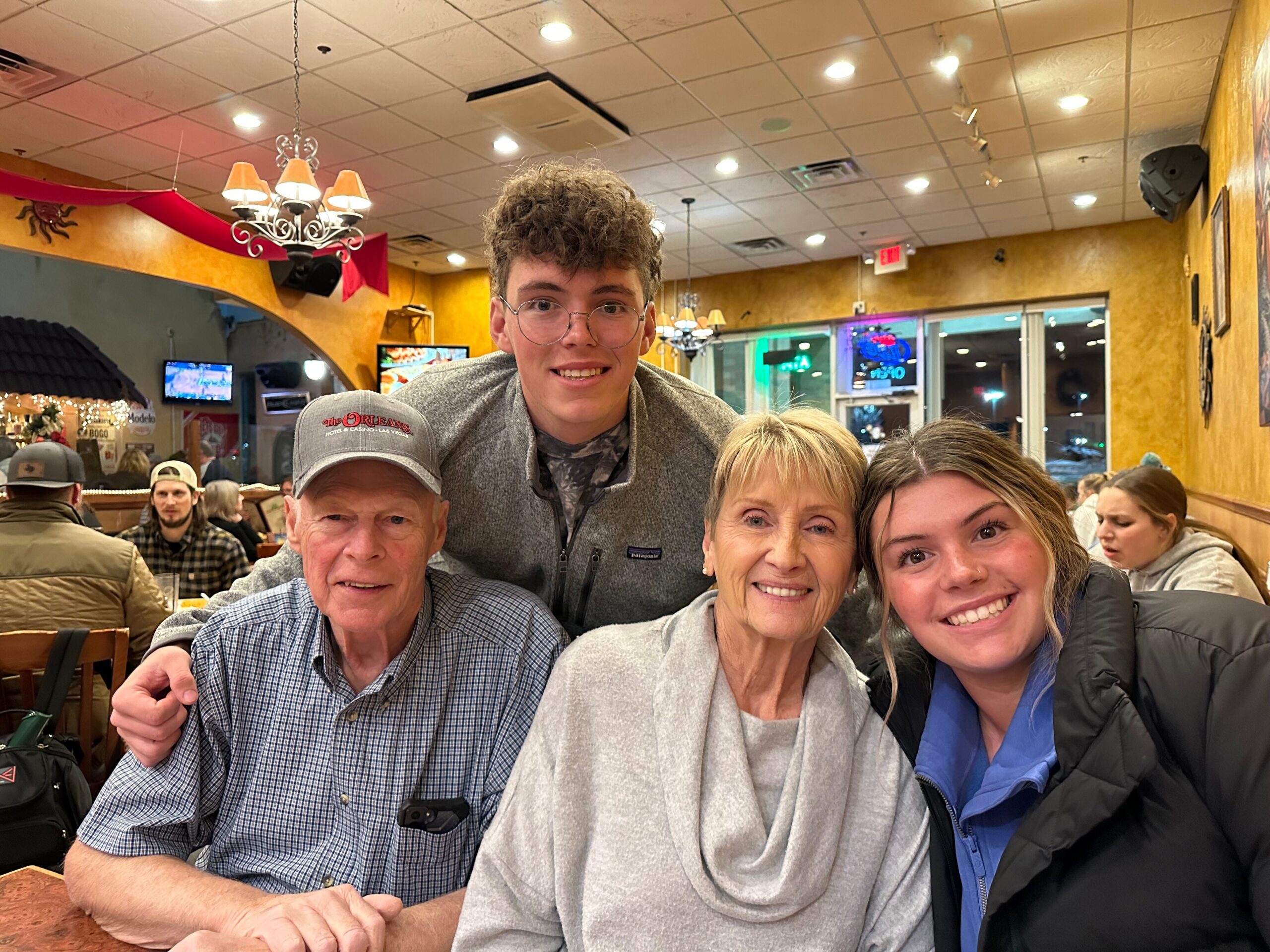 Group of five friends/family posing together in a crowded restaurant, smiling at camera under warm lighting.