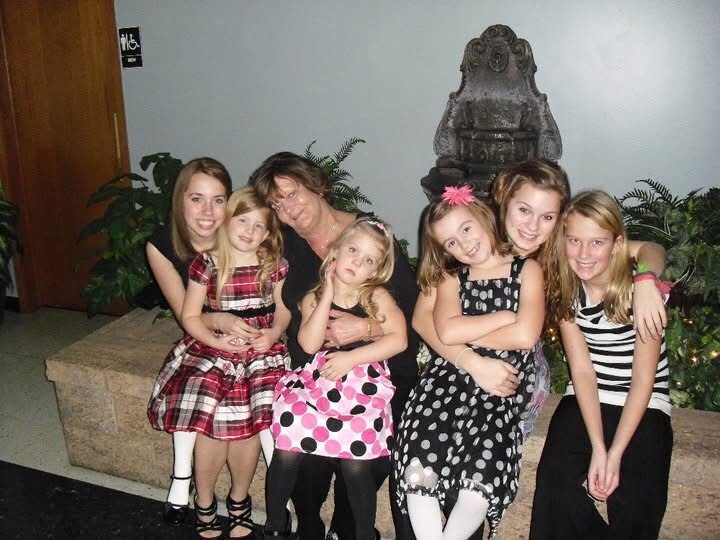 A woman sits with six young girls in colorful dresses, posing together near a decorative indoor fountain in a lobby-like space by plants.
