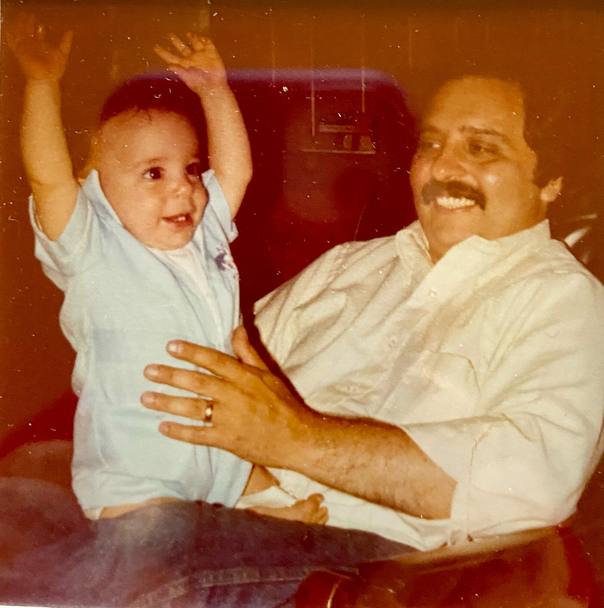 Man with a mustache smiling as he holds a laughing baby who raises both arms, seated in a car with a headrest visible behind them (sepia photo).