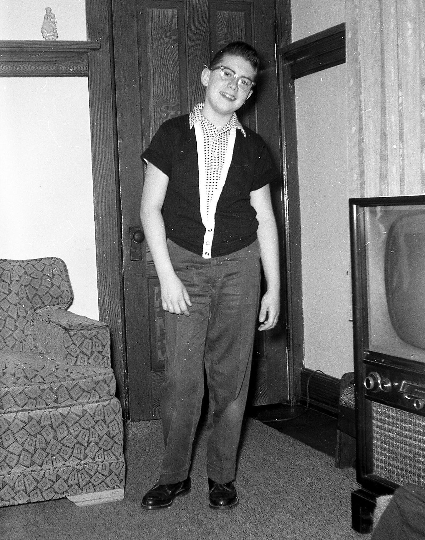 Teenager with glasses in a doorway of a retro living room, wearing a dotted shirt and dark cardigan, with a vintage TV to the right.