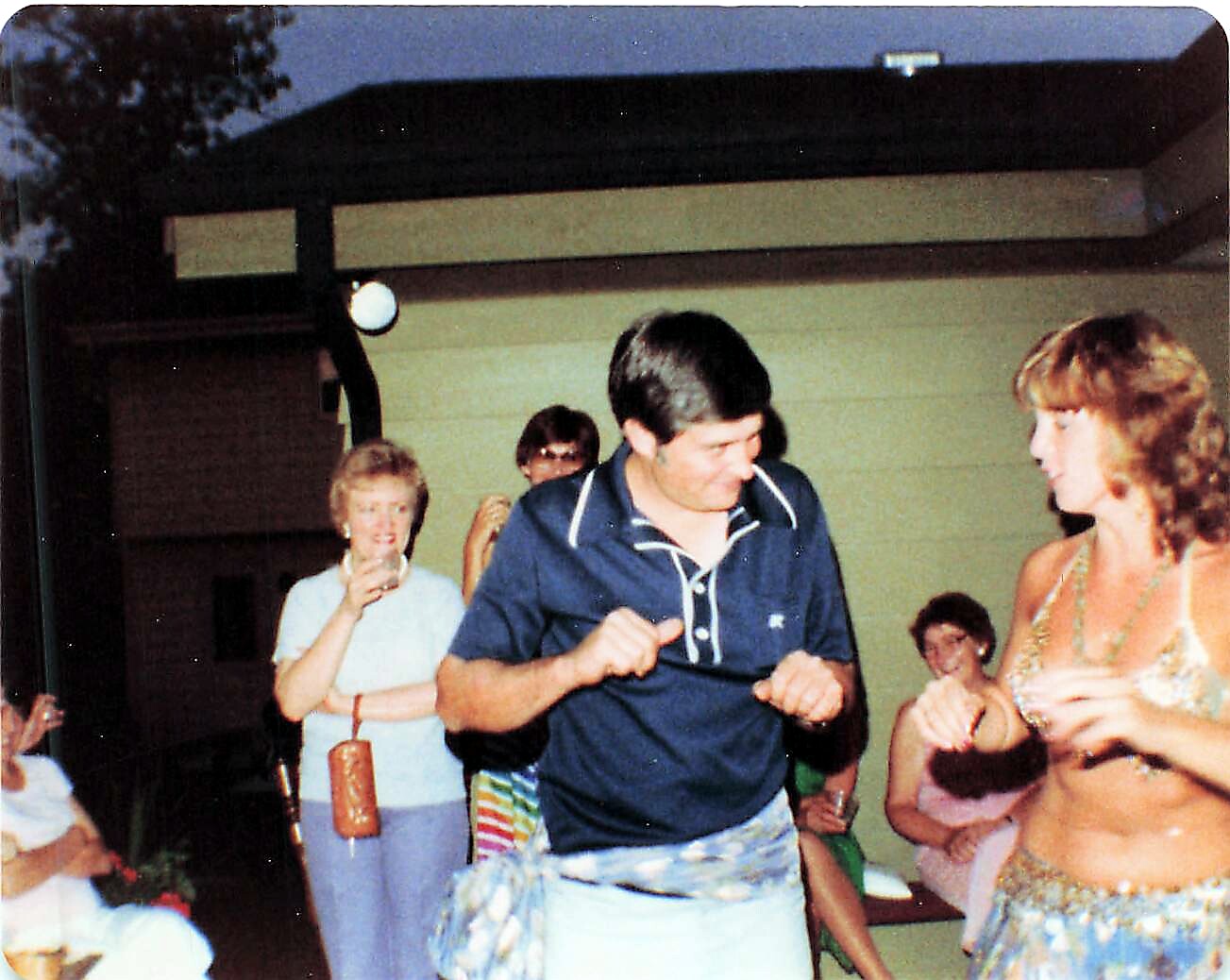 Man in a navy polo dances with a belly-dancer as friends watch at an outdoor party.