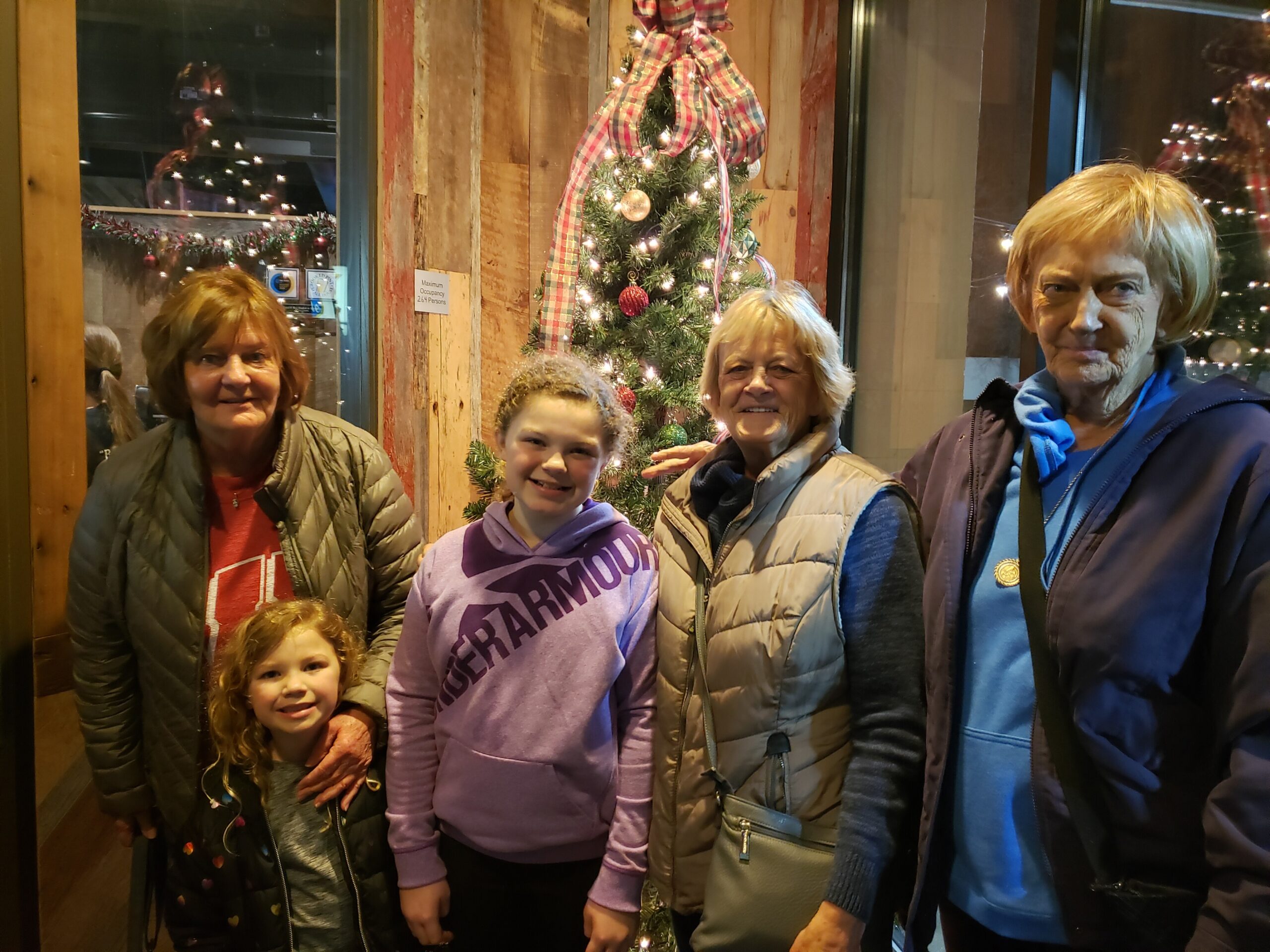 Five people (two children in front, three adults behind) posing indoors beside a lit Christmas tree with plaid ribbon decoration.