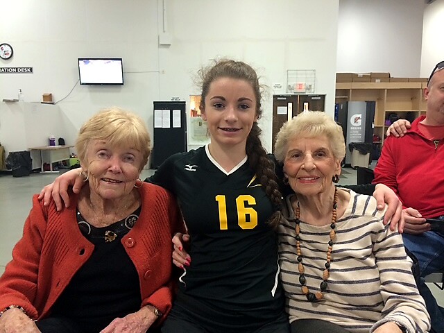 Young female athlete in a black jersey with the number 16 sits between two smiling elderly women in a light-colored room, arms around each other, posing for a photo in a gym or recreation center.