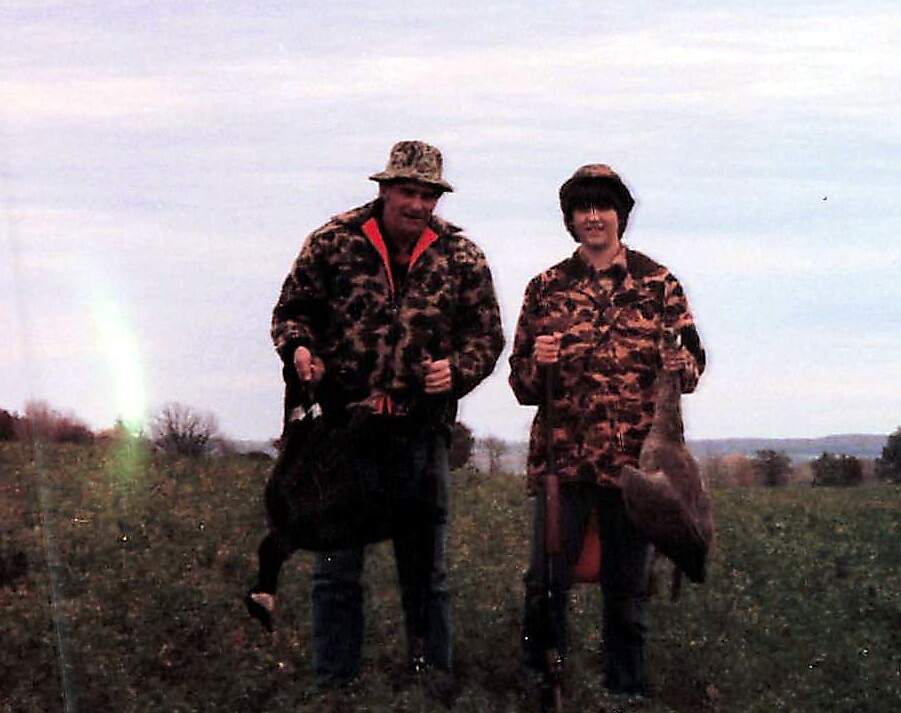 Two hunters in camouflage jackets stand in an open field, posing with game birds and carrying bags