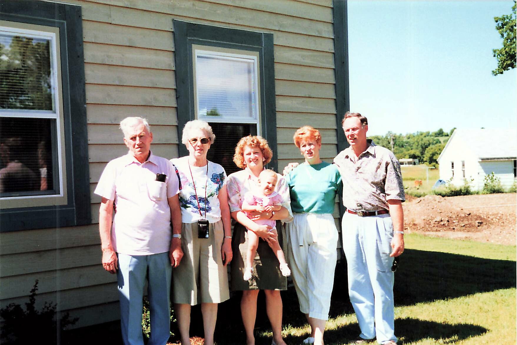 Group of six adults and a baby posing together outside a beige wooden house on a sunny day.