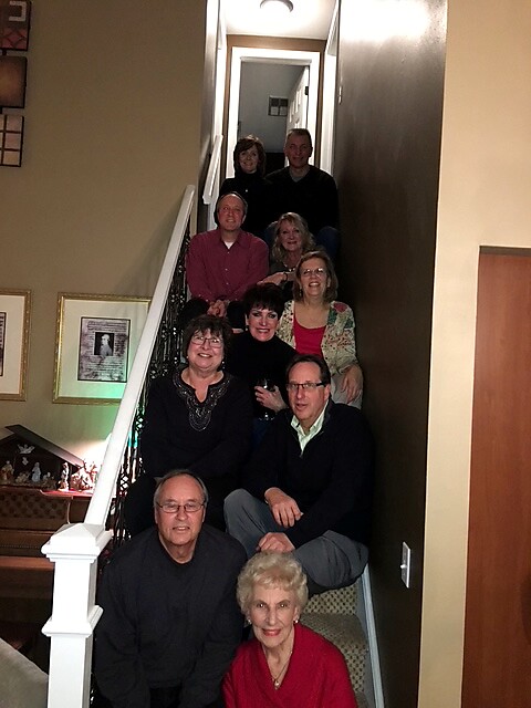 Group of twelve adults posing on a carpeted staircase inside a home, all smiling for a casual portrait.