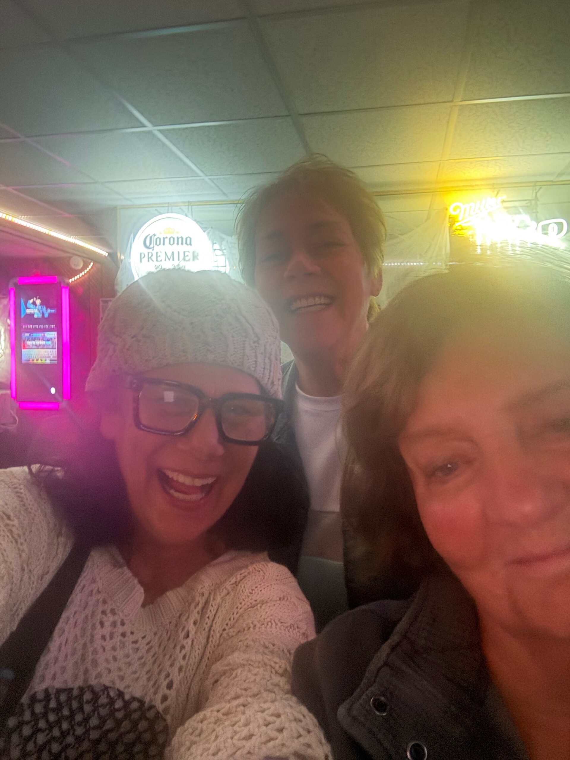 Group selfie of three friends smiling in a neon-lit bar, with a Corona Premier sign in the background, wearing sweaters and jackets.