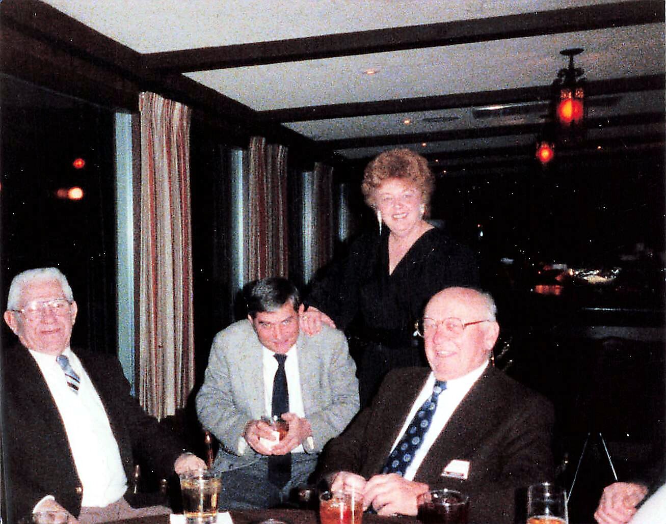 Four people at a dinner table in a dim restaurant: three men seated with drinks, and a smiling woman standing behind them.