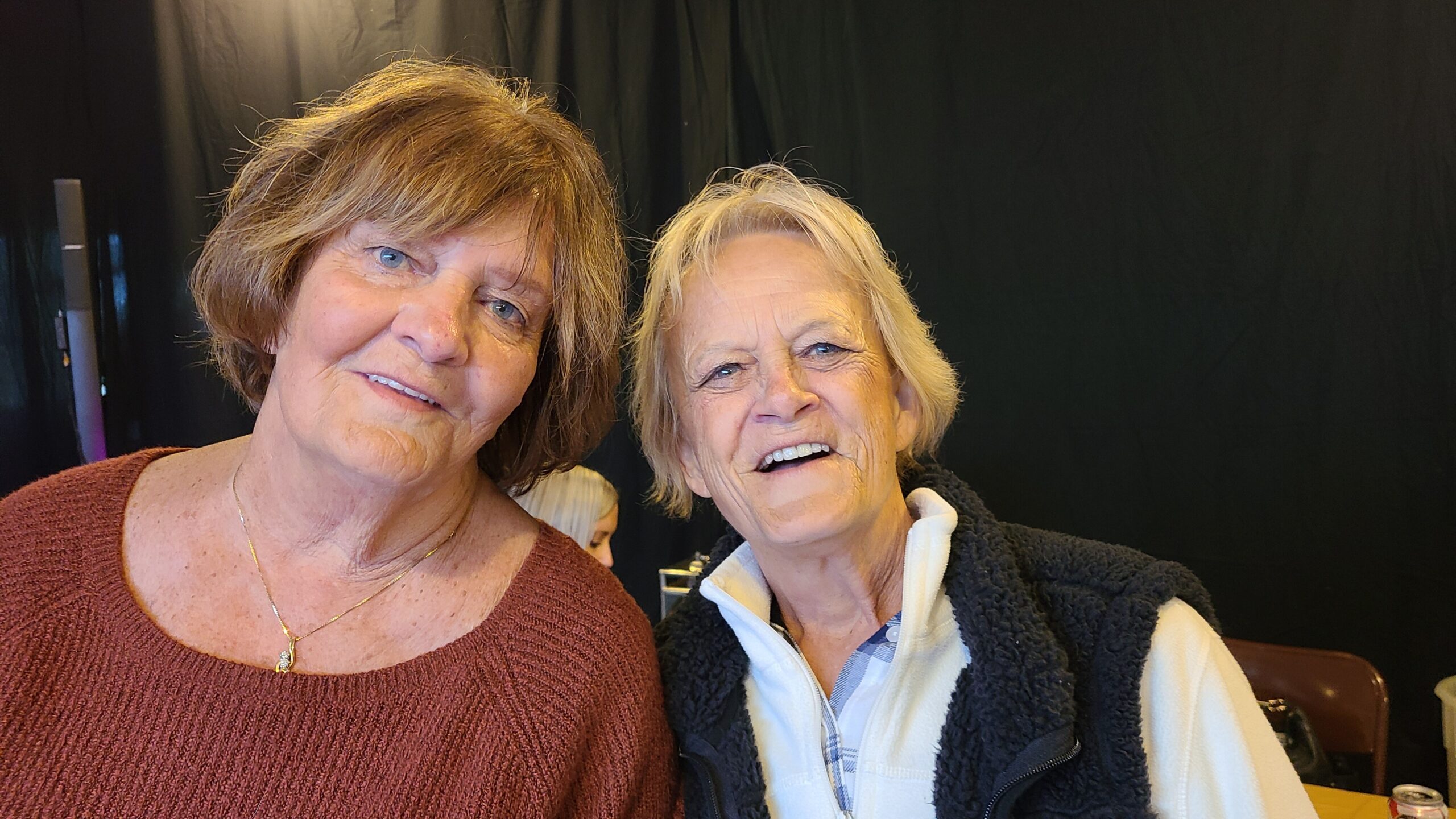 Two elderly women smiling at the camera in a casual indoor setting, one with short brown hair wearing a rust-colored sweater and a gold necklace, the other with light blonde hair in a white fleece vest.