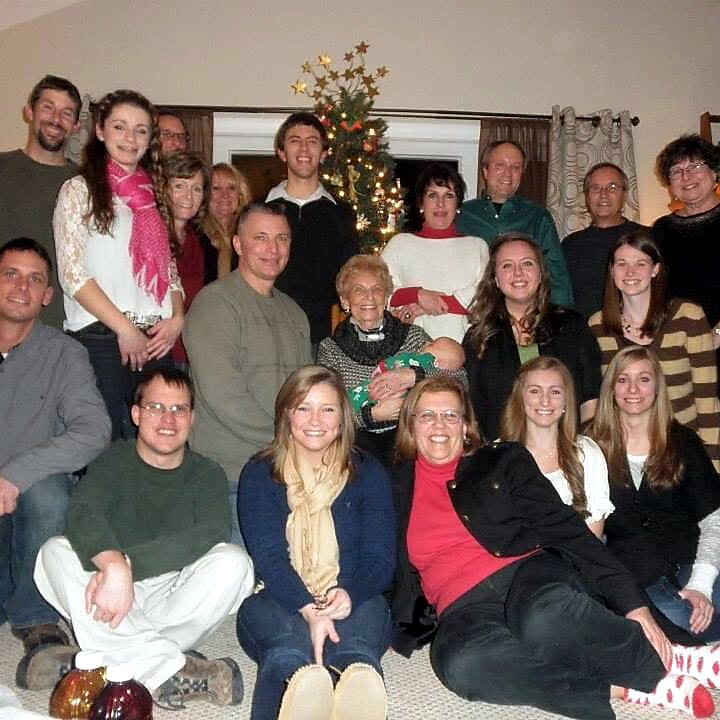 Group of about 20 people posing for a Christmas portrait in a living room, with a lit Christmas tree in the background and everyone smiling.