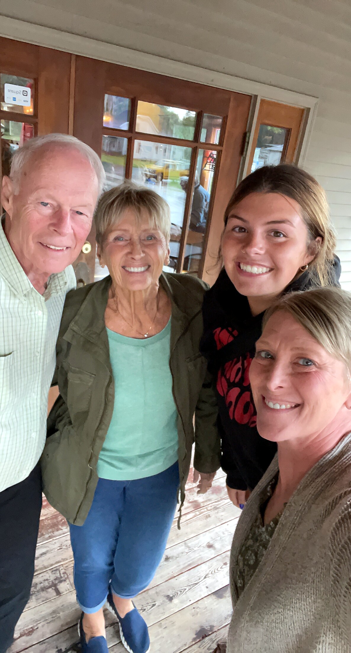 Four adults posing for a selfie on a wooden porch in front of glass-pane doors, smiling at the camera.