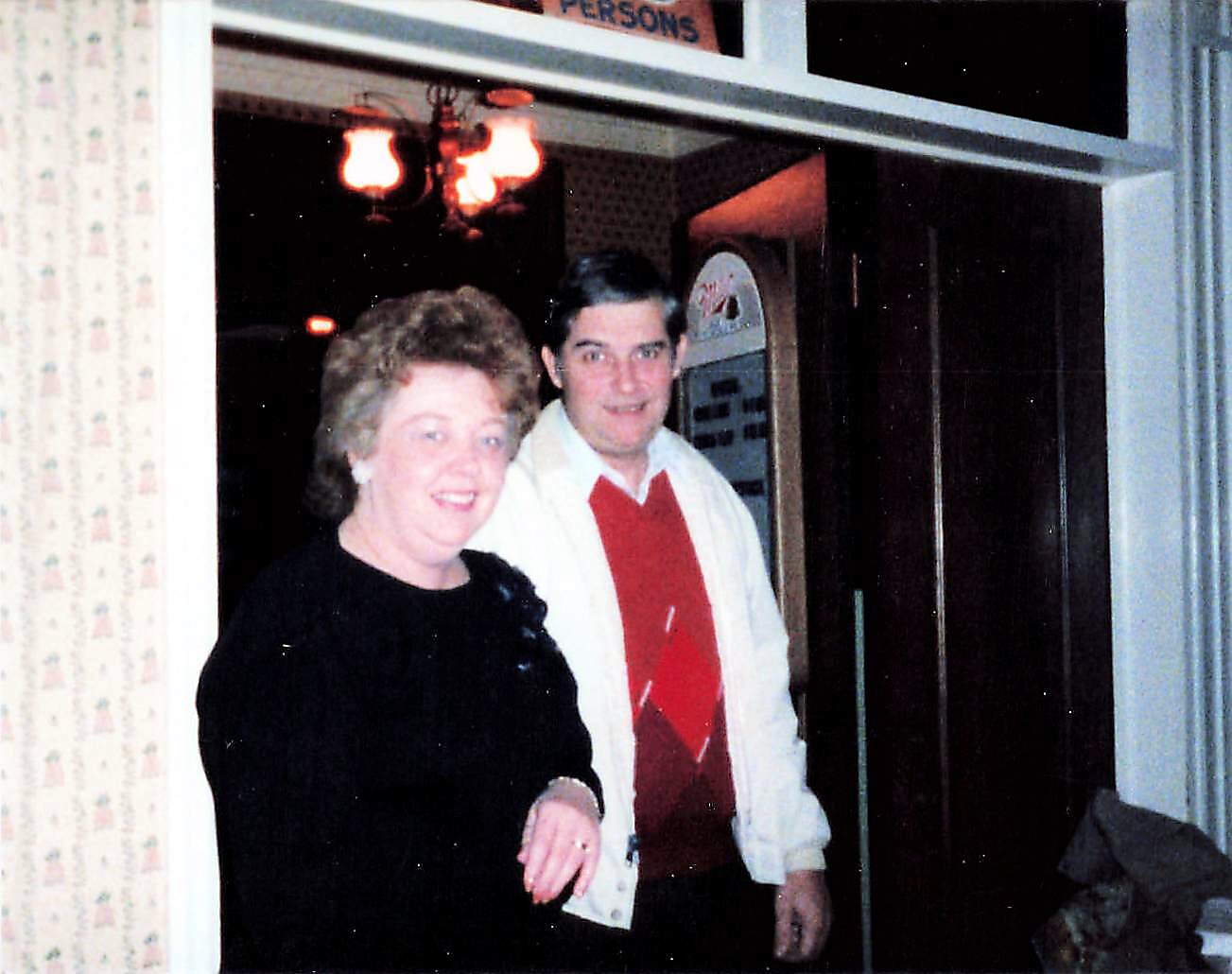 Smiling woman in a black dress and a man in a white jacket and red sweater pose in a doorway inside a diner with a jukebox visible behind them.