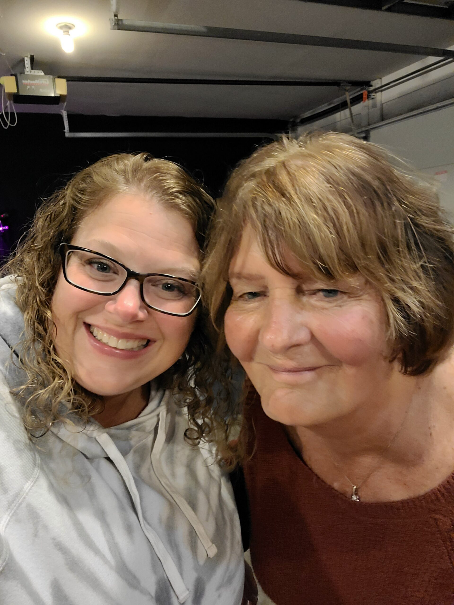 Two women smiling and leaning close for a selfie indoors; one wears glasses and a light gray hoodie, the other in a rust-colored top.