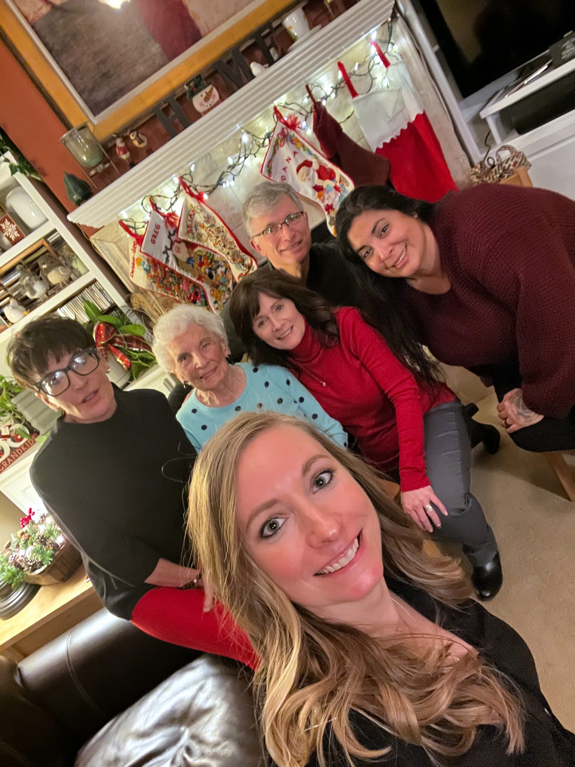 Group of smiling adults posing for a Christmas selfie in a festively decorated living room with stockings and lights.