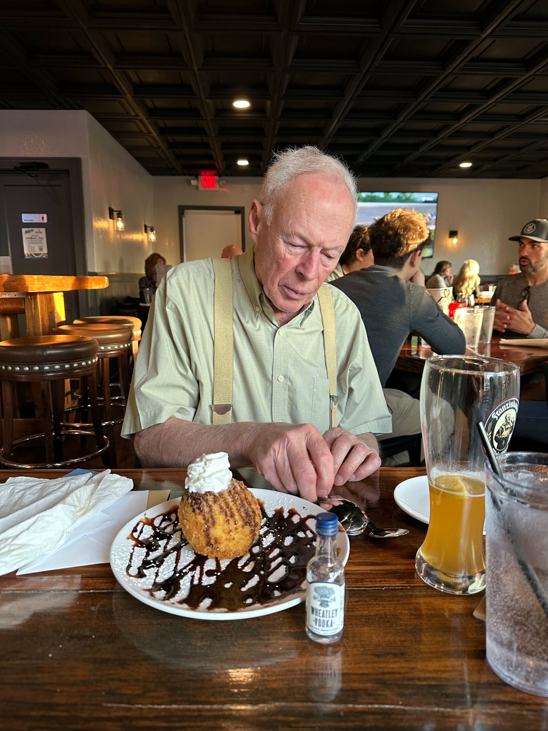 Older man in a light green shirt and suspenders sits at a wooden table, looking down at a plated dessert with chocolate drizzle and a dollop of whipped cream; a beer glass and small vodka bottle are nearby in a busy restaurant setting.