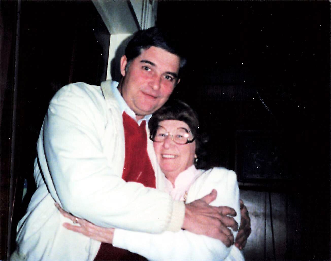 A man in a white jacket hugs an older woman in a light pink blouse and glasses, both smiling at the camera.