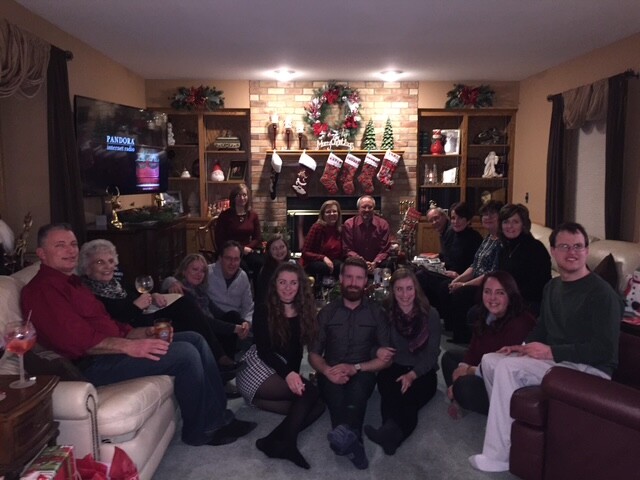 A large group of family and friends gathers in a cozy living room for a Christmas party, seated and kneeling around a central coffee table in front of a brick fireplace with stockings and holiday decorations.