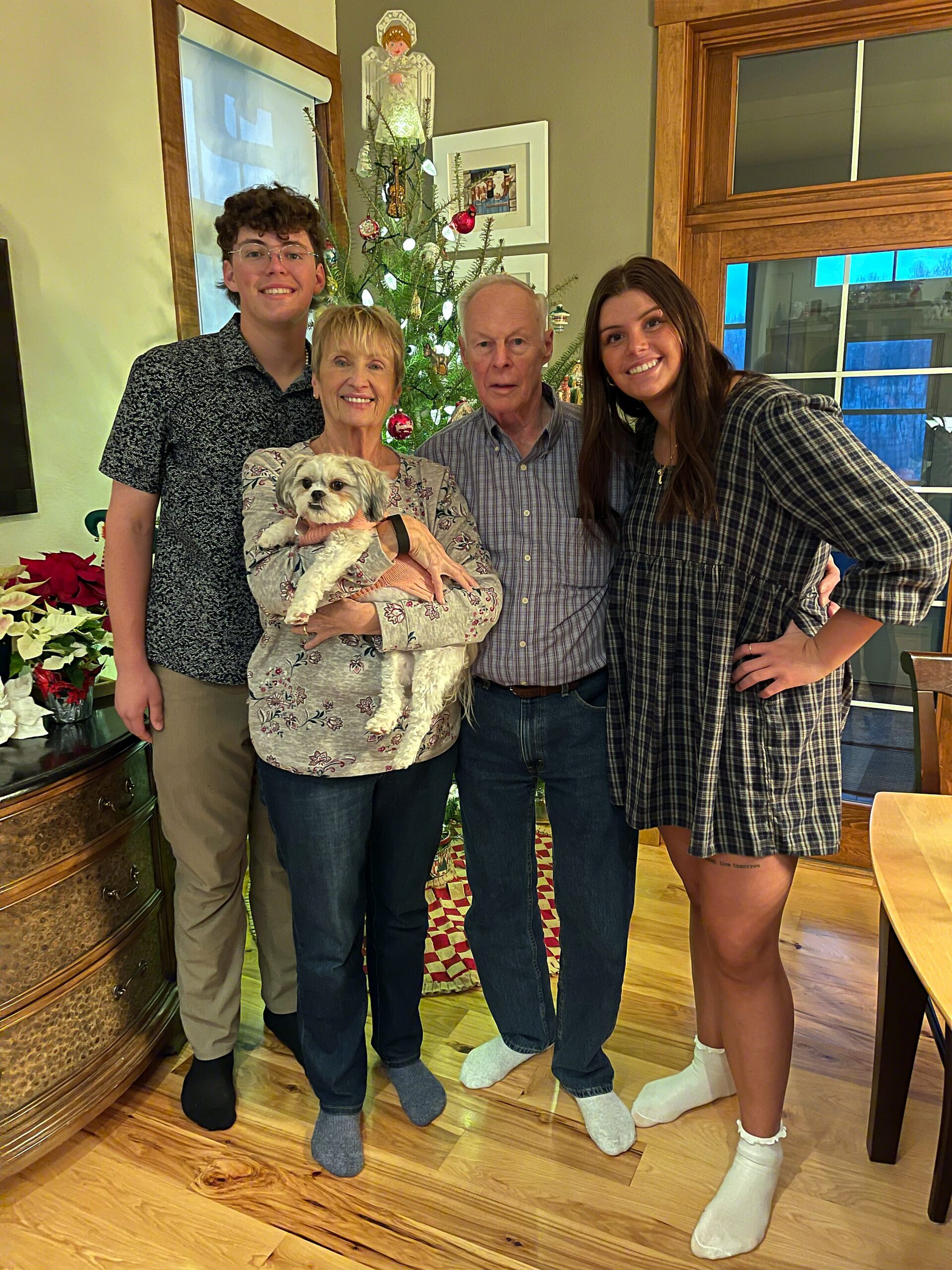 A family group posing indoors by a Christmas tree: older couple with a small dog, and two young adults smiling at the camera.