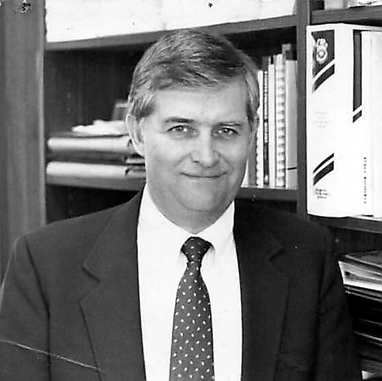 Smiling man in a suit and tie seated in an office with bookshelves behind him.