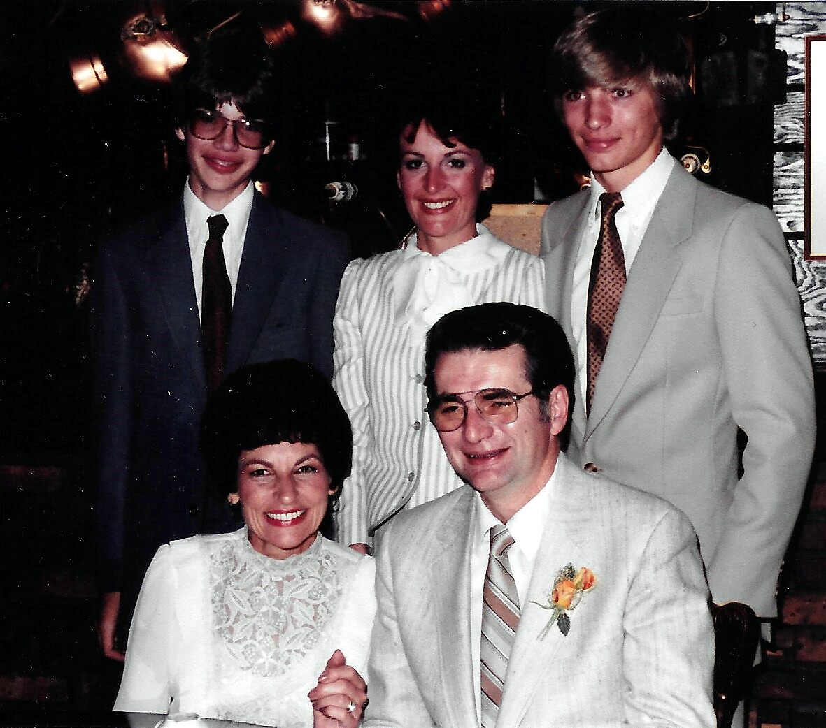 Smiling couple seated front at a formal event, woman in a white lace blouse and man in a light suit with a boutonniere, with two young adults behind them.